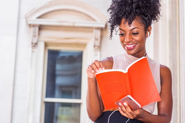 Young Happy African American female college student with afro hairstyle sitting by vintage style office building in New York, reading red book, listening music with earphone and cell phone, smiling.