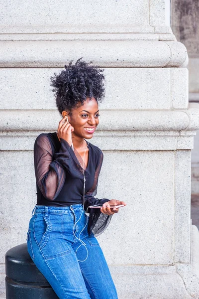 Young African American Woman with afro hairstyle, wearing mesh sheer long sleeve shirt blouse, blue jeans, sitting on street in New York City, listening music with earphone and cell phone, texting