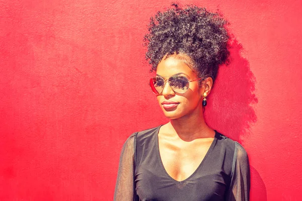 New York'ta Genç Afro-Amerikan Kadın Portresi. Uzun kollu örgü sırf gömlek giyen afro saç modeli ile genç siyah kadın, güneş altında kırmızı arka plan karşı duran, güneş gözlüğü
