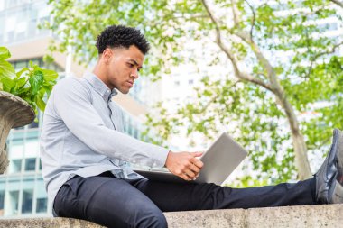 New York'ta okuyan genç Amerikan Koleji Öğrencisi, açık gri uzun kollu gömlek giyiyor, siyah pantolon, deri ayakkabı, kampüste yeşil ağaç tarafından duvarın üstünde oturan, dizüstü bilgisayar üzerinde çalışan