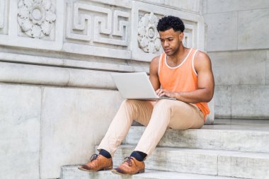 New York'ta okuyan genç Mix-Race Amerikan üniversite öğrencisi, turuncu tank üst giyiyor, bej pantolon, kahverengi deri ayakkabı, kampüste ofis dışında merdivenlerde oturan, dizüstü bilgisayar üzerinde çalışan