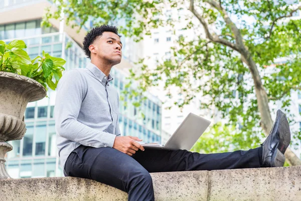 Young American College Student studying in New York City, wearing light gray shirt, black pants, leather shoes, sitting on top of wall by green tree on campus, working on laptop computer, thinking