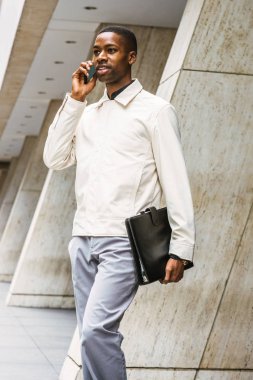 Genç Afro-Amerikan işadamı seyahat, New York'ta çalışan, Genç siyah adam açık renkli ceket giyiyor, deri çanta taşıyan, ofis binasından dışarı yürüyüş, cep telefonu konuşuyor