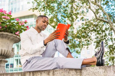 Genç Afro-Amerikan Adam dışarıda kitap okuyor. Açık renkli ceket giyen genç siyah adam, gri pantolon, deri ayakkabı, Manhattan sokak sokak yarım duvarın üstünde oturan, New York, kırmızı kitap okuma