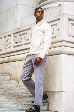 Genç Yakışıklı Afro-Amerikan Adam açık renkli ceket giyiyor, gri pantolon, siyah deri ayakkabı, New York kampüsünde vintage mermer duvar ile merdivenlerde duran, etrafında dönüyor, uzağa bakıyor