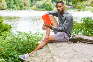 Genç Afro-Amerikan Adam New York Central Park'ta rahatlatıcı. Gri gömlek, şort, spor ayakkabı giyen, göl kenarında ki kayalıklarda oturan, kitap okuyan genç siyah adam. Backgroun'da kürekli insanlar