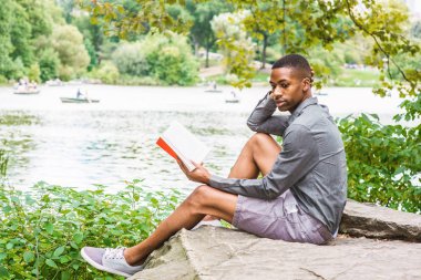 Genç Afro-Amerikan Adam New York Central Park'ta rahatlatıcı. Gri gömlek, şort, spor ayakkabı giyen, göl kenarında ki kayalıklarda oturan, kitap okuyan genç siyah adam. Backgroun'da kürekli insanlar