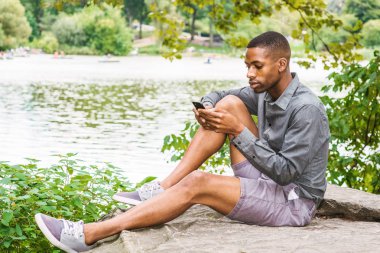 Genç Afro-Amerikan Adam New York Central Park'ta rahatlatıcı. Gri uzun kollu gömlek, şort, spor ayakkabı giyen genç siyah adam, göl kenarında kayalarda oturuyor, okuyor, cep telefonuyla mesaj atıyor.
