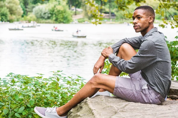 Genç Afro-Amerikan Adam New York Central Park'ta rahatlatıcı. Uzun kollu gri gömlek, şort giyen, göl kenarında ki kayalıklarda oturan genç siyah adam, düşünüyor. Birçok kişi arka planda tekne kürek