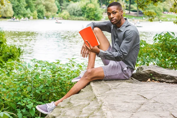 Genç Afro-Amerikan Adam New York Central Park'ta rahatlatıcı. Gri gömlek, şort, spor ayakkabı giyen, göl kenarında ki kayalıklarda oturan, kitap okuyan genç siyah adam. Backgroun'da kürekli insanlar