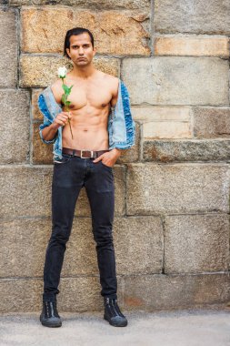 Genç Doğu Hint Amerikan Man mavi moda Denim ceket giyen, açıldı, yarı çıplak güçlü vücut gösteren, siyah kot pantolon, spor ayakkabı, beyaz gül tutan, New York'ta kaya duvarının yanında duran, bekliyor