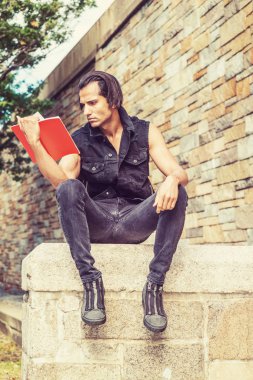 Genç Modern Doğu Hint Amerikan Man siyah kolsuz ceket giyiyor, kot pantolon, spor ayakkabı, New York'ta parkta taş duvar taş çit üzerinde oturan, aşağı bakıyor, kırmızı kitap okuma