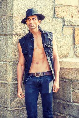 Genç Doğu Hint Amerikan Man siyah kolsuz moda Denim ceket giyiyor, düğmesiz, yarı çıplak vücut gösteren, kot pantolon, Fedora şapka, New York'ta kaya duvar ve çit tarafından ayakta, uzağa bakıyor
