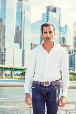 Genç Doğu Hint Amerikan Man beyaz gömlek giyiyor, siyah kot pantolon, New York Hudson Nehri tarafından yüksek binalar ile iş bölgesinde ayakta, ileriye bakıyor
