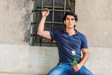 Seni özlüyorum ve seni düşünüyorum. Genç Adam mavi kısa kollu gömlek giyiyor, kot pantolon, New York'ta sokakta pencere ile duvara karşı oturan, beyaz gül çiçeği tutan, ciddi ileriye bakıyor