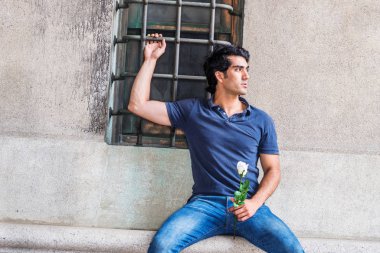 Seni özlüyorum ve seni düşünüyorum. Genç Adam mavi kısa kollu gömlek giyiyor, kot pantolon, New York'ta sokakta pencere ile duvara karşı oturan, beyaz gül çiçeği tutan, ciddi uzağa bakıyor