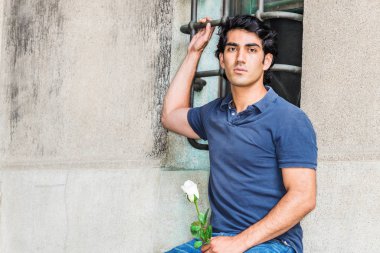 Seni özlüyorum ve seni düşünüyorum. Genç Adam mavi kısa kollu gömlek giyiyor, kot pantolon, New York'ta sokakta pencere ile duvara karşı oturan, beyaz gül çiçeği tutan, ciddi ileriye bakıyor