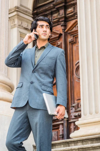 Young Mix Race American Businessman working in New York City, wearing dark blue jacket, pants, holding laptop computer, walking dow stairs from old style office building, calling on cell phone