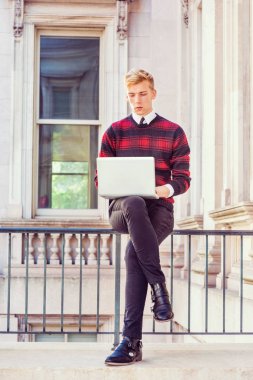 Genç Amerikalı üniversite öğrencisi okuyor, New York'ta çalışıyor, desenli kırmızı, siyah örgü kazak, siyah pantolon, deri ayakkabı giyiyor, ofis binasında korkulukta oturuyor, dizüstü bilgisayarda çalışıyor.