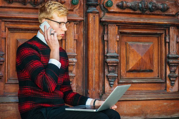 Young  blonde American college student wearing patterned red, black knit sweater, glasses, sitting by brown vintage wooden office door, working on laptop computer, talking on cell phone