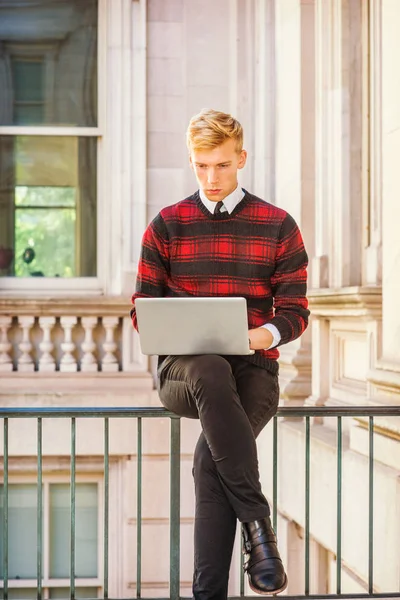 Young blonde American college student wearing patterned red, black knit sweater, black pants, leather shoes, sitting on railing inside office building, looking down, working on laptop computer