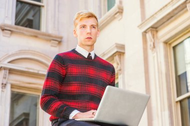 Genç sarışın Amerikan üniversite öğrencisi desenli kırmızı giyiyor, siyah örgü kazak, kampüste vintage ofis binası dışında oturan, dizüstü bilgisayar üzerinde çalışan, yukarı bakıyor, düşünme