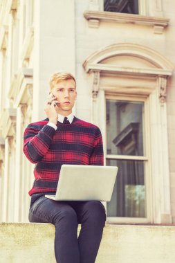Genç sarışın Amerikalı adam desenli kırmızı giyiyor, siyah örgü kazak, kampüste vintage ofis binası dışında oturan, dizüstü bilgisayarüzerinde çalışan, cep telefonu konuşuyor, düşünme