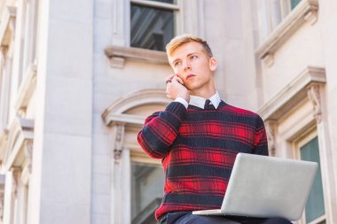 Genç sarışın Amerikalı adam desenli kırmızı giyiyor, siyah örgü kazak, kampüste vintage ofis binası dışında oturan, dizüstü bilgisayar üzerinde çalışan, cep telefonu konuşuyor