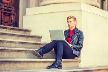 Genç sarışın Amerikan Adam okuyor, New York'ta çalışıyor, siyah deri ceket, pantolon, deri ayakkabı giyiyor, kampüsteofiyer binasında merdivenlerde oturuyor, okuyor, dizüstü bilgisayarda çalışıyor