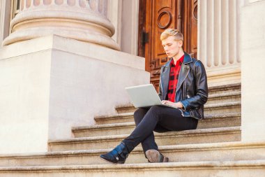 Genç sarışın Amerikan Adam okuyor, New York'ta çalışıyor, siyah deri ceket, pantolon, deri ayakkabı giyiyor, kampüsteofiyer binasında merdivenlerde oturuyor, okuyor, dizüstü bilgisayarda çalışıyor