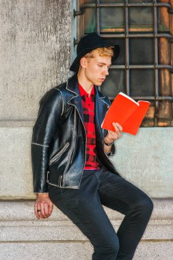 Başarıya Giden Yol. New York'ta okuyan genç Amerikan Adam, siyah deri ceket, kırmızı desenli gömlek, siyah şapka giyiyor, sokakta penceresi olan vintage duvara karşı oturuyor, kırmızı kitap okuyor