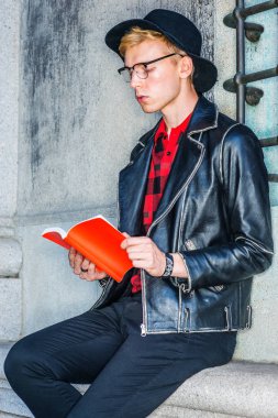 Başarıya Giden Yol. New York'ta okuyan genç Amerikalı üniversite öğrencisi, siyah deri ceket, kırmızı desenli gömlek, siyah şapka, gözlük giyiyor, kampüste vintage duvara karşı oturuyor, kırmızı kitap okuyor