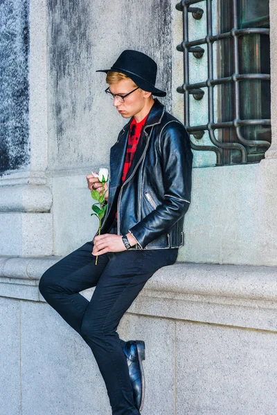 Missing You. Young American Man wearing black leather jacket, glasses, hat, sitting by vintage wall on street in New York, looking down at white rose, hand touching petal, sad, thinking