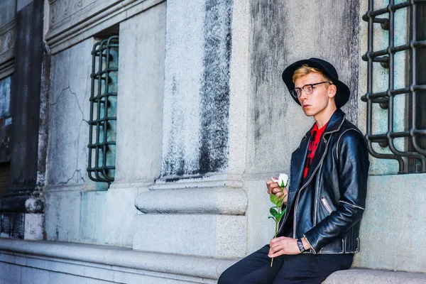 Missing You. Young American Man wearing black leather jacket, glasses, hat, sitting by vintage wall on street in New York, holding white rose, hand touching petal, looking away, frowned, sad, thinking