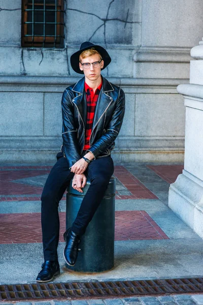 Young Man Casual Spring/Autumn Fashion in New York, wearing black leather jacket, patterned red undershirt, pants, leather shoes, hat, glasses, sitting on metal pillar on vintage street, relaxing