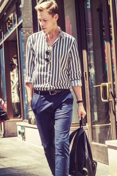 Young European Man traveling in New York City, wearing black, white striped, long sleeve shirt, blue pants, hand holding bag, walking out from store in Midtown of Manhattan, looking down, think.