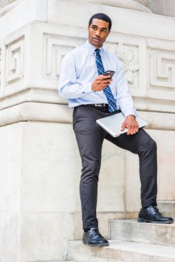 Genç Afro-Amerikan İşadamı seyahat, New York'ta çalışan, beyaz gömlek, kravat, siyah pantolon giyen, dizüstü bilgisayar tutan, sokakta mermer duvar tarafından merdivenlerde duran, cep telefonuyla mesajlaşma