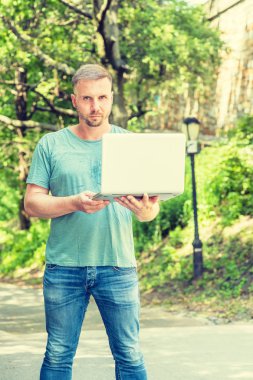 Sakallı genç Amerikalı Adam, New York'ta çalışıyor, açık yeşil tişört giyiyor, kot pantolon giyiyor, dizüstü bilgisayar tutuyor, okul kampüsünde yeşil ağaçlarla sokakta duruyor, okuyor, düşünüyor.