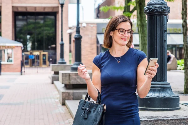 Young 40 years old Native American Teacher working in New York, wearing short sleeve dress, glasses, arm carrying bag, standing on street by light poles on school campus, reading messages on phone