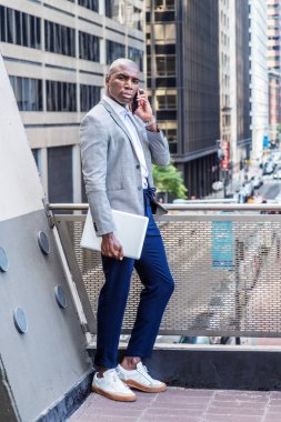 Gri desenli spor ceket, mavi pantolon, beyaz spor ayakkabı, dizüstü bilgisayar, balkonda korkuluk başında bekleyen genç Afrikalı Amerikalı adam, yüksek binaların olduğu sokağa bakıyor, cep telefonuyla konuşuyor.