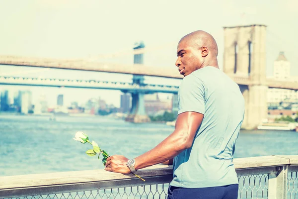 Welcome You. I miss you. Young African American Man wearing gray T shirt. holding white rose, standing by fence at harbor in New York City, waiting for you. Manhattan, Brooklyn bridges on background.