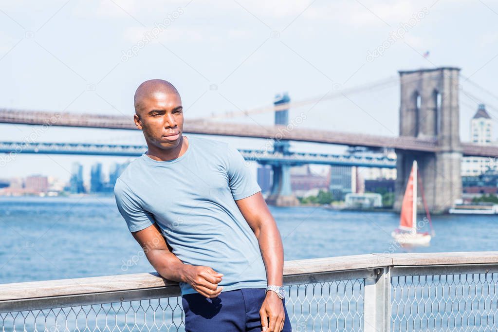 I missing you, waiting for you. Young African American Man traveling in New York City, wearing gray T shirt, standing by East River, looking forward. Manhattan, Brooklyn bridges on background