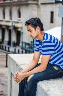 Yalnız Doğu Hintli Amerikalı genç New York 'un dışında düşünüyor, mavi çizgili tişört giyiyor, modası geçmiş kot pantolon giyiyor, sokakta oturuyor, üzgün, depresif, düşüncelere dalmış
