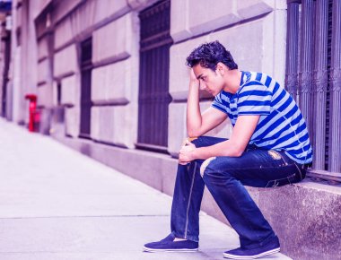 New York 'un dışında, mavi çizgili tişört, yırtık kotlar, kumaş ayakkabılar giymiş, büyük bir pencerenin çerçevesinde oturan, el ele tutuşan üzgün bir Amerikalı genç.