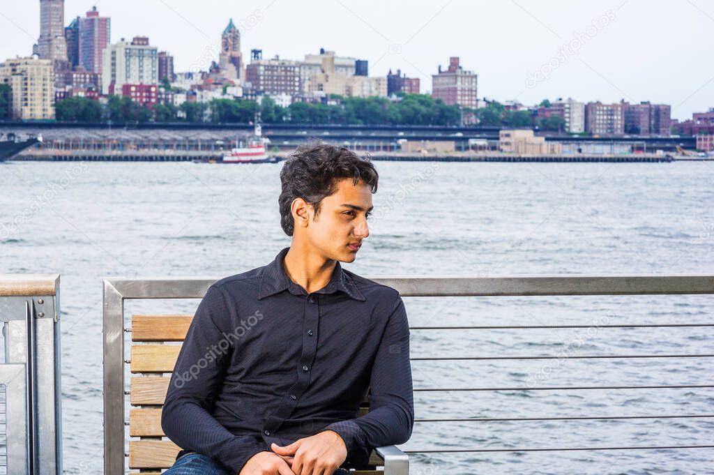 18 years old East Indian American male teenager relaxing outside in New York City, wearing black long sleeve shirt, sitting on chair by East River, looking around, thinking