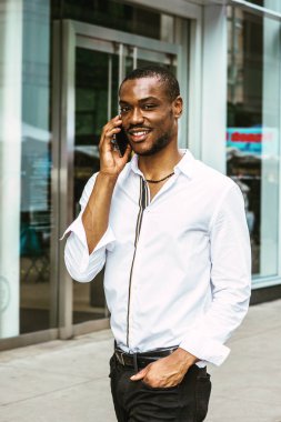 New York 'ta sakallı, beyaz uzun kollu gömlekli, siyah pantolonlu, Manhattan' da modern ofis binasının önünden geçen, cep telefonuyla konuşan, gülümseyen genç bir Afrikalı Amerikalı..