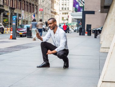 Sakallı, beyaz uzun kollu gömlekli, siyah pantolonlu, New York 'taki ofis binasının önünde oturan, cep telefonuyla okuyan, düşünen genç bir Afrikalı.