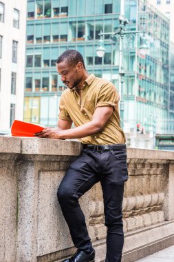 Sakallı, kısa kollu yeşil gömlekli, siyah pantolonlu, Manhattan, New York 'taki ofis binasının önünde duran, kırmızı kitap okuyan genç bir Afrikalı Amerikalı.