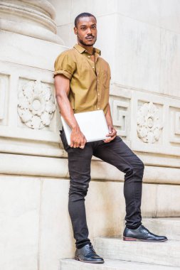 New York 'ta sakallı, yeşil kısa kollu gömlekli, siyah pantolonlu, deri ayakkabılı, dizüstü bilgisayar taşıyan, beyaz mermer duvarda duran Afrikalı genç bir üniversite öğrencisi.