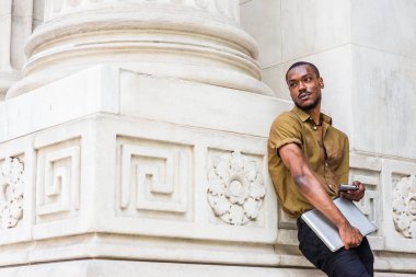 New York 'ta çalışan genç Afrikalı Amerikalı iş adamı, yeşil kısa kollu tişört giyiyor, dizüstü bilgisayar taşıyor, ofis binasının önünde duruyor, cep telefonuyla mesajlaşıyor, başka tarafa bakıyor, düşünüyor.